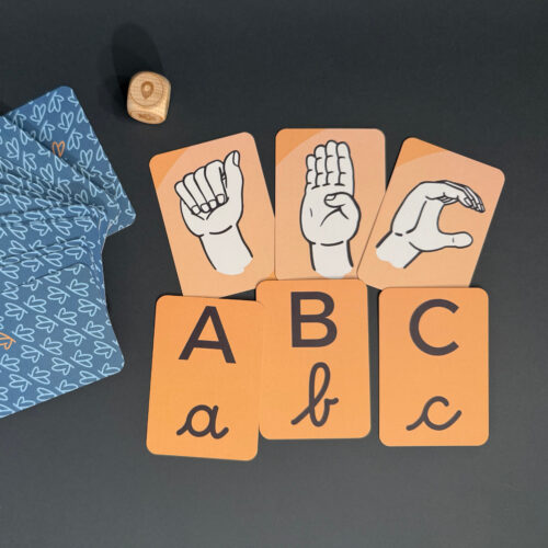 Ozadje je temno sivo, karte so v črkah ABC in v ročni abecedi. Zraven je kocka.