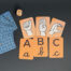 Ozadje je temno sivo, karte so v črkah ABC in v ročni abecedi. Zraven je kocka.