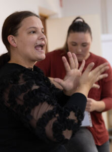 Jerneja uporablja znakovni jezik. V ozadju Daša gleda v vrečko. 